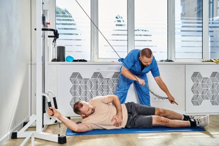 Photo for Young doctor in blue uniform assisting man training on exercise machine in gym of kinesio center - Royalty Free Image