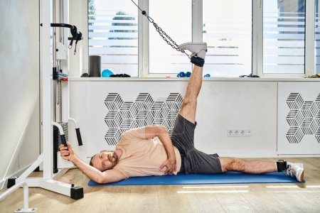 hombre en ropa deportiva acostado en la alfombra de fitness y la pierna de entrenamiento en la máquina de ejercicio en el centro kinesio