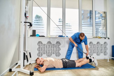 Photo for Physician in blue uniform assisting man working out on exercise machine in gym of kinesio center - Royalty Free Image