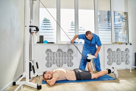 Photo for Physician in blue uniform assisting man working out on exercise machine in modern kinesio center - Royalty Free Image