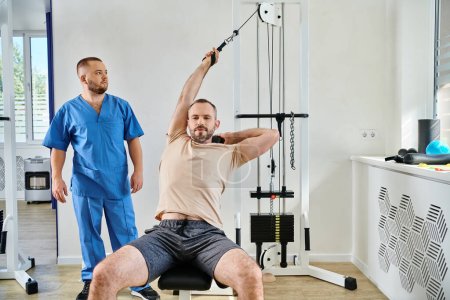 man in sportswear working out on exercise machine near professional assistant in kinesio center