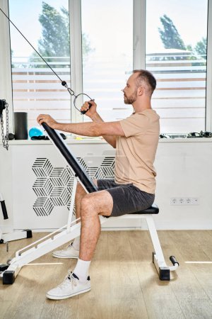 Photo for Side view of handsome bearded man in sportswear working out on training machine in kinesio center - Royalty Free Image