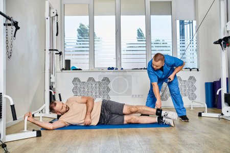 Photo for Young specialist in blue uniform assisting male patient during recovery training in kinesio center - Royalty Free Image