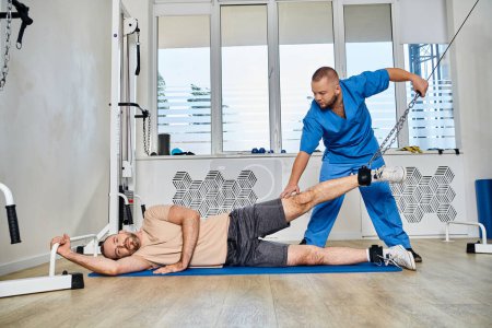 Photo for Skilled rehabilitologist in blue uniform assisting man during recovery training in kinesio center - Royalty Free Image