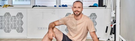 Photo for Joyful bearded man in sportswear sitting at exercise machine in contemporary kinesio center, banner - Royalty Free Image