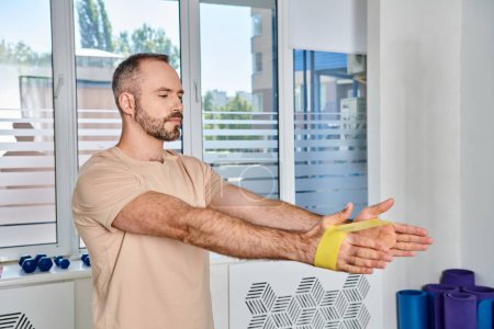 Photo for Handsome bearded man training with resistance band in kinesiology center, recovery session - Royalty Free Image