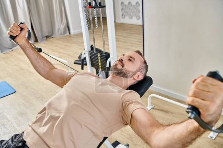 Photo for Bearded man in sportswear training arms on exercise machine in kinesio center, recovery session - Royalty Free Image