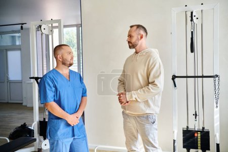 bearded man and professional rehabilitologist in blue uniform talking in modern kinesio center