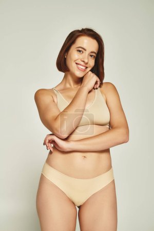 body positive, young woman in underwear smiling and posing with hand near face on grey background