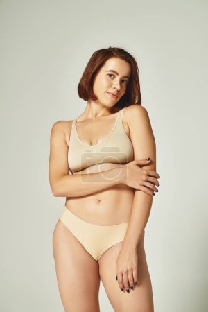 body positive, pretty woman in underwear smiling and posing with hand near body on grey background