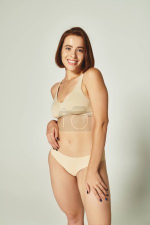 body positive, cheerful young woman with short hair standing in beige underwear on grey background