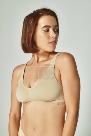 self-assured and young woman in comfortable beige lingerie looking away on grey background
