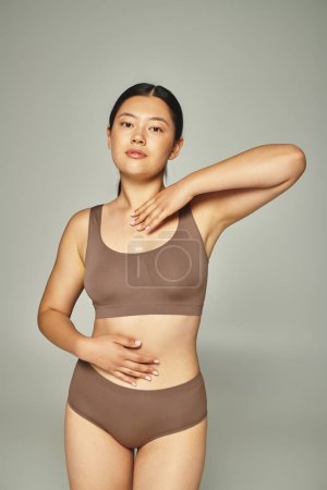 young asian woman with brunette hair posing in brown underwear on neutral grey background