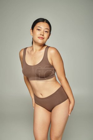 charming young asian woman with brunette hair posing in brown lingerie on grey background