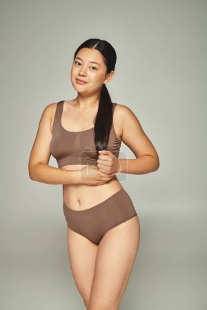 charming young asian woman with shiny brunette hair posing in brown lingerie on grey background
