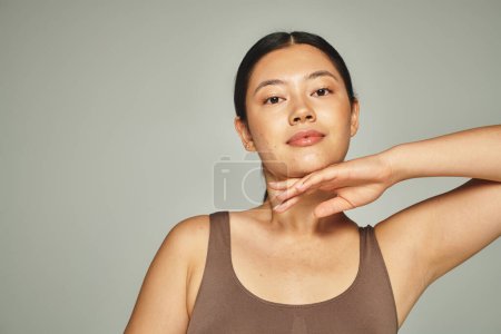pretty asian woman in brown top touching her face on grey background, skin care and beauty