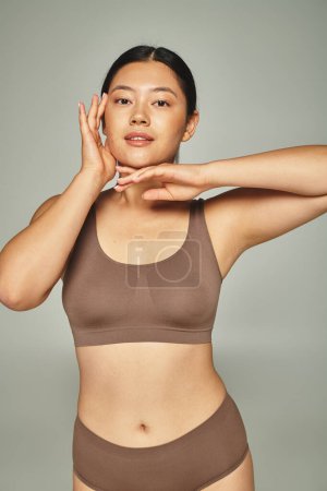 brunette asian woman in brown top touching her face on grey background, skin care and beauty