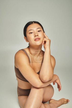 Téléchargez les photos : Brunette jeune femme asiatique en sous-vêtements assis avec les jambes croisées sur fond gris, corps positif - en image libre de droit