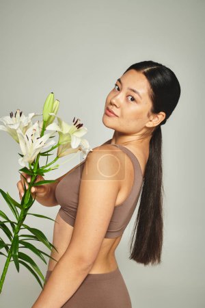 Téléchargez les photos : Brunette asiatique femme en pastel sous-vêtements tenant des fleurs et debout sur fond gris, lis - en image libre de droit