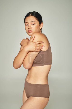 schüchterne asiatische Frau in Unterwäsche, die ihren Körper bedeckt, während sie sich auf grauem Hintergrund umarmt, body shaming