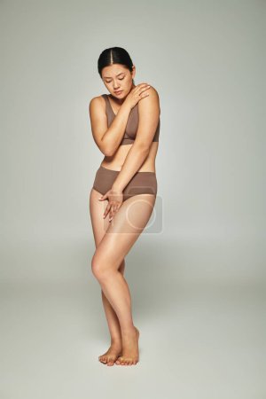 aufgebrachte asiatische Frau in Unterwäsche, die ihren Körper bedeckt, während sie sich vor grauem Hintergrund umarmt, Body Shaming