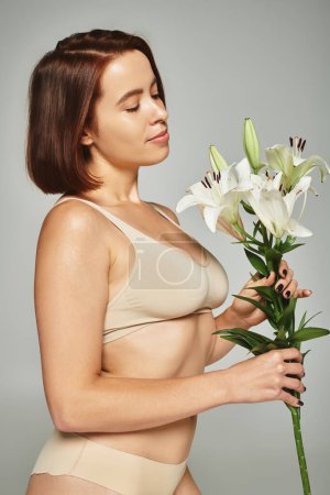young woman with short hair holding flowers and posing in underwear on grey background, lilies