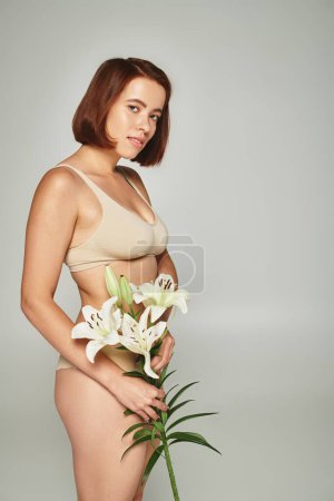 charming woman with short hair holding flowers and posing in underwear on grey background, lilies