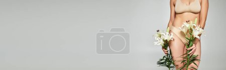 femme en lingerie beige tenant des lys blancs et debout sur fond gris, bannière recadrée