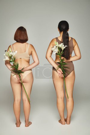 Rückansicht zweier Frauen in beiger Dessous, die mit weißen Lilien in der Hand vor grauem Hintergrund stehen