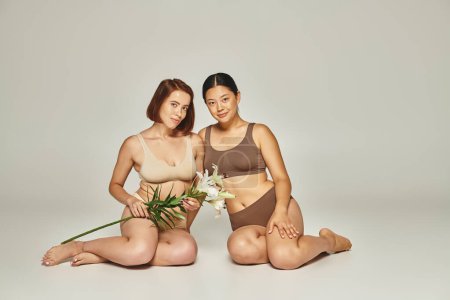 young multicultural women in underwear sitting together with flowers on grey background, lilies