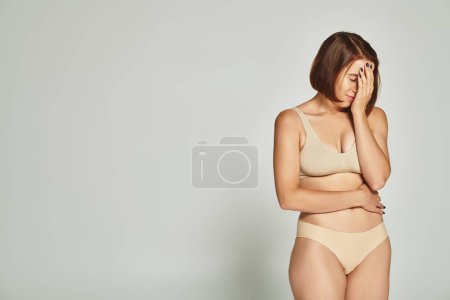 embarrassed young woman in beige underwear covering face with hand on grey background, body shaming