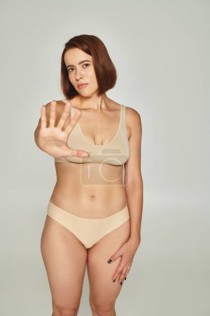 young woman in beige underwear gesturing and showing stop on grey background, body shaming