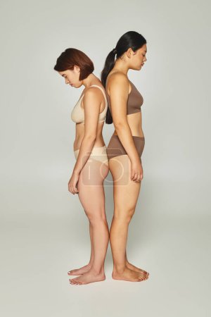 Aufgebrachte multikulturelle Frauen in Unterwäsche, die Rücken an Rücken vor grauem Hintergrund stehen, Body Shaming