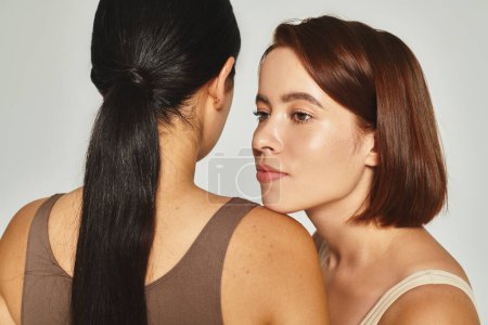 young woman with short hair standing near brunette female friend on grey background, body positive