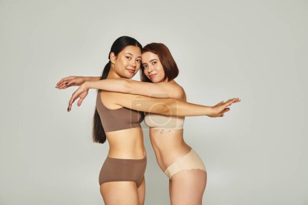 happy woman embracing with asian female friend in underwear while posing together on grey backdrop