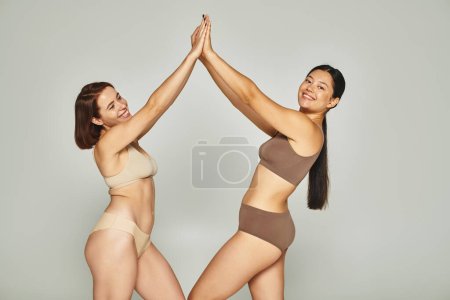 junge glückliche interrassische Frauen in Unterwäsche High-Fiving einander auf grauem Hintergrund, Körper positiv