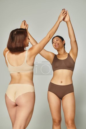 young happy interracial women in lingerie high-fiving each other on grey background, body positive