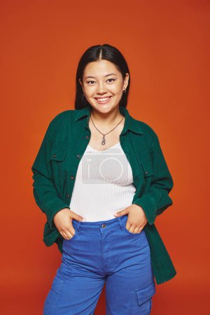 cheerful young asian woman posing in vibrant outfit with hands in pockets on orange background