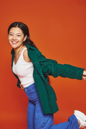 happy young asian woman posing in vibrant outfit with outstretched hands on orange background