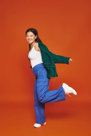 joyful young asian woman posing in vibrant outfit with outstretched hands on orange background