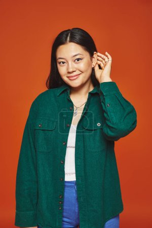 joyful young asian woman posing in vibrant outfit and adjusting brunette hair on orange background