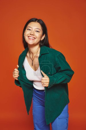radiante joven asiático chica posando en vibrante traje y usando verde chaqueta en naranja fondo