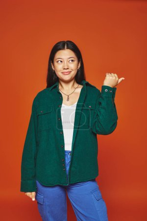 Photo for Asian woman posing in vibrant outfit with green jacket pointing away with thumb on orange background - Royalty Free Image