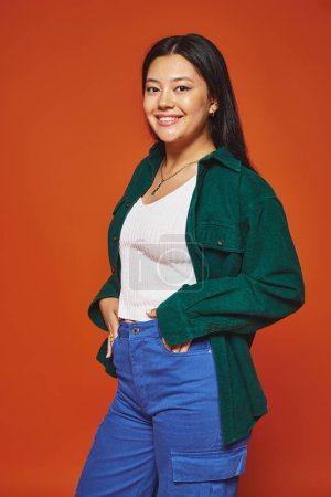 happy young asian woman posing in vibrant outfit with hands in pockets on orange background