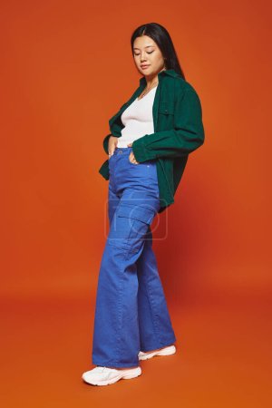 stylish young asian woman posing in vibrant outfit with hands in pockets on orange background