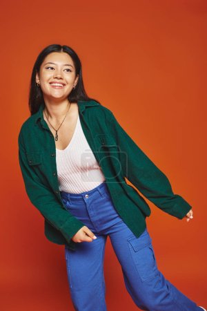 cheerful young asian woman posing in vibrant outfit and looking away on orange background