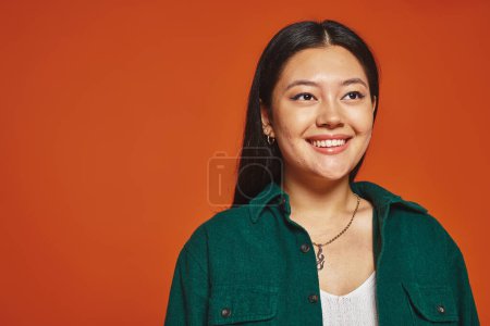 Porträt einer strahlenden und glücklichen asiatischen Frau in grünem Sakko, die auf orangefarbenem Hintergrund lächelt,