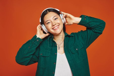 junge und brünette asiatische Frau genießt Musik mit drahtlosen Kopfhörern auf orangefarbenem Hintergrund
