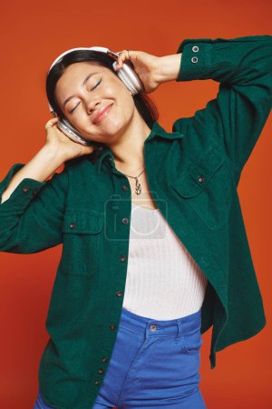 pleased brunette asian woman enjoying music with wireless headphones on orange background