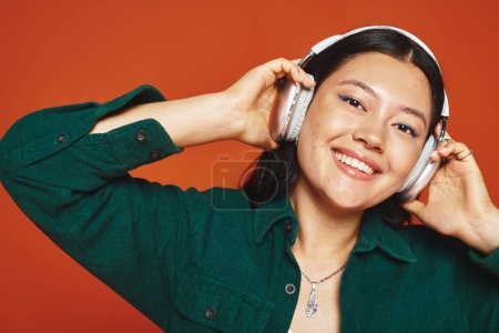 alegre morena asiática mujer disfrutando de la música con auriculares inalámbricos sobre fondo naranja
